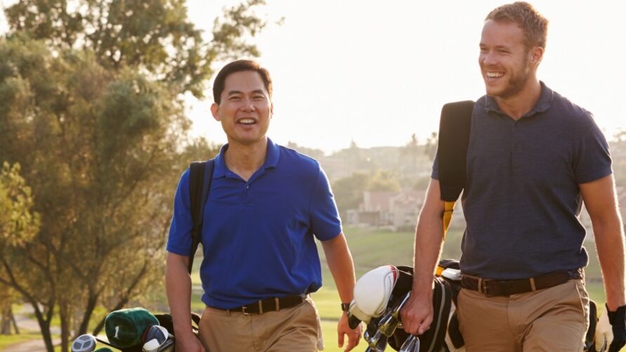 two male friends walk across the golf course carrying their clubs