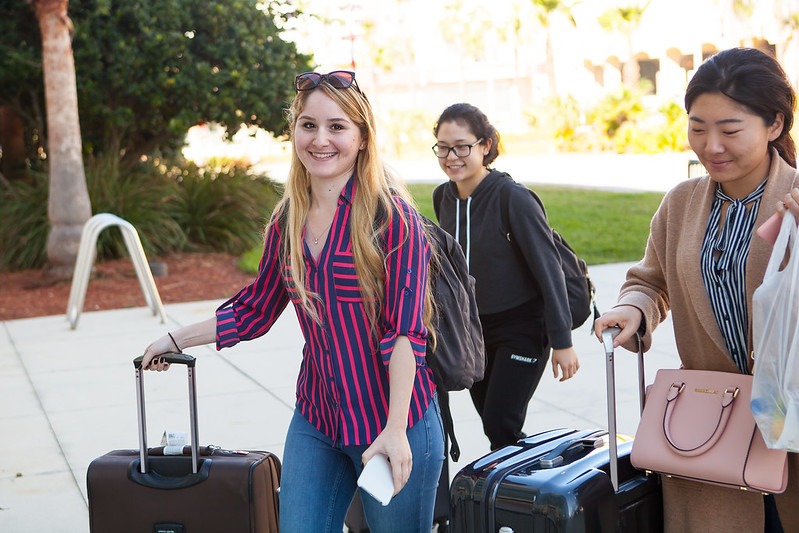 3 female international students pulling large suitcases walk together outside
