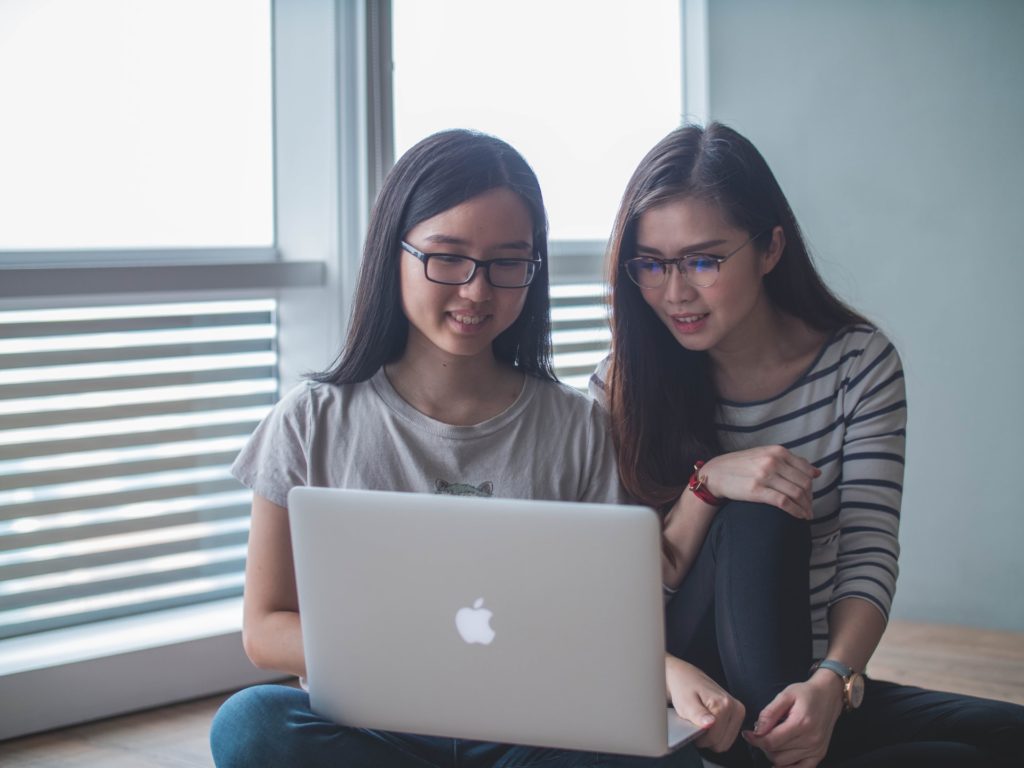 two asian girls sit in a window seat looking at a computer together