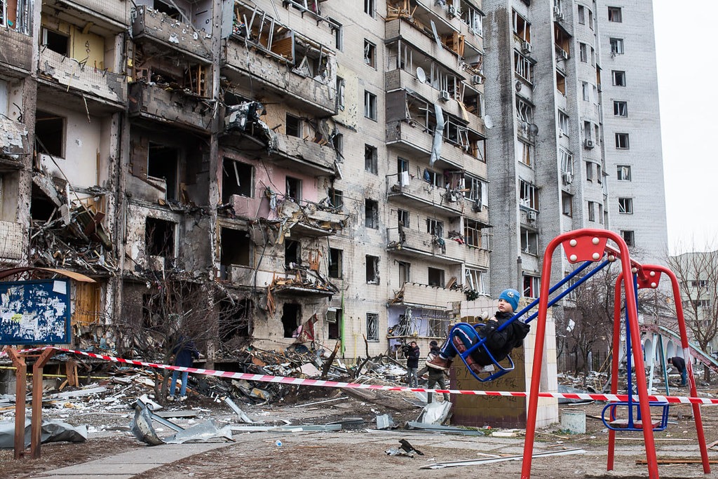 young boy swinging on a playground in front of a destroyed building