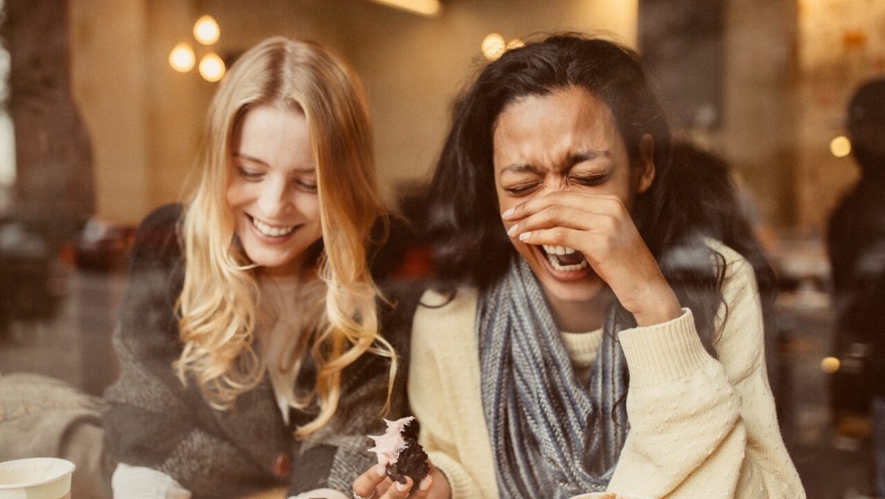 two female friends laugh together while enjoying cupcakes and coffee in a cafe