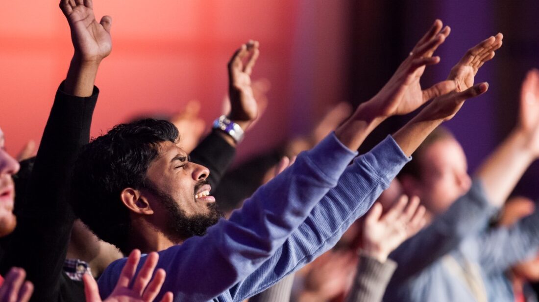 south asian man sings worship songs with arms raised in a large group